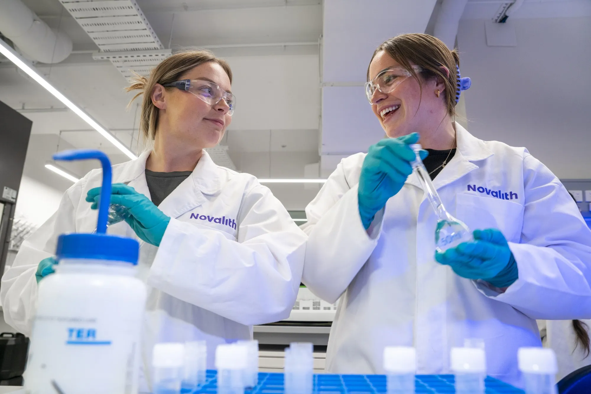 Two Novalith scientists in lab coats and safety glasses, smiling as one holds up a flask of sample in the Sydney laboratory