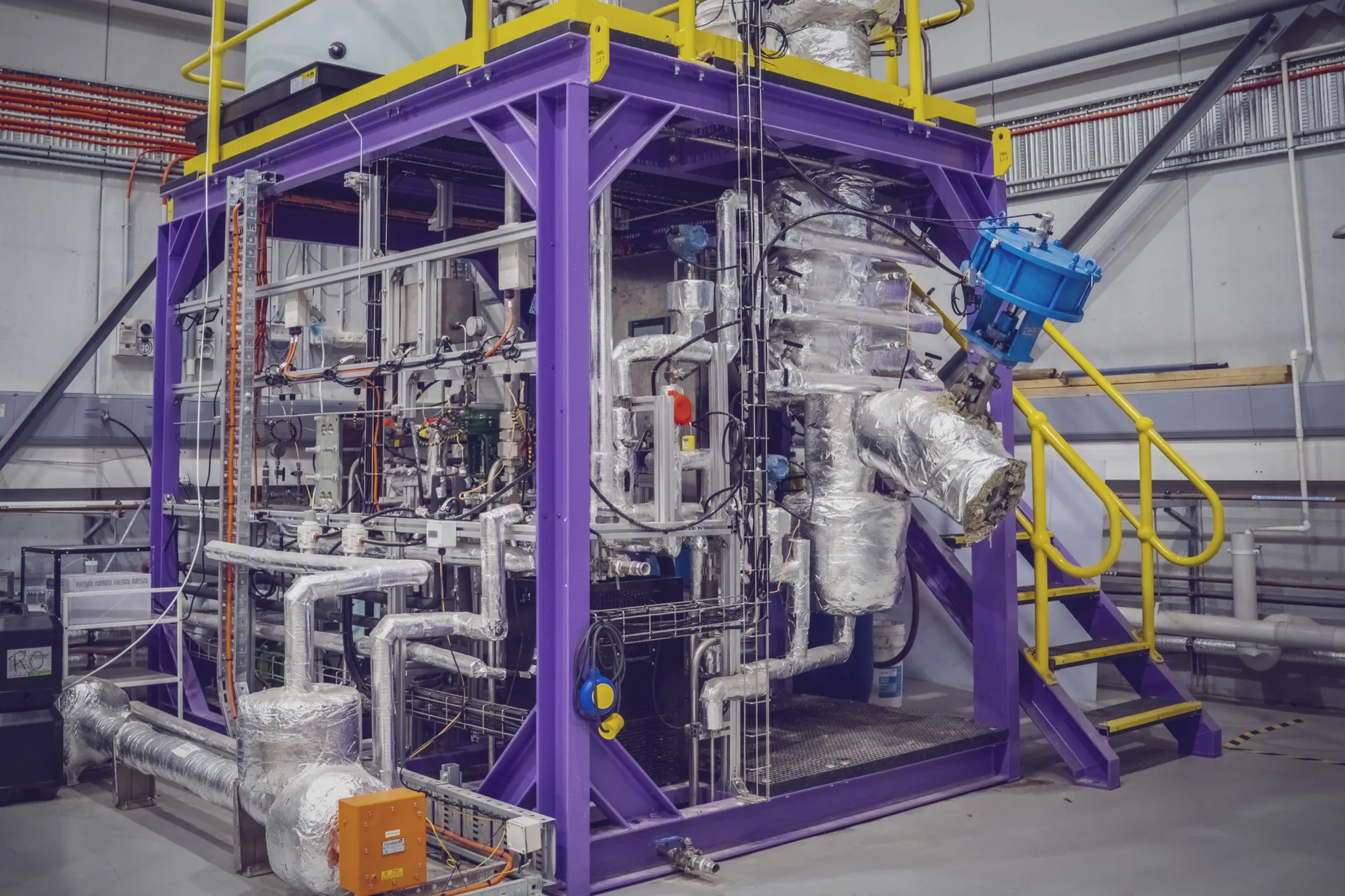 The Novalith pilot plant in Sydney: a two-storey purple steel frame carrying the insulated reactor and pipework, with a yellow handrail around the upper platform.